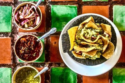 Guacamole servi dans un molcajete de pierre avec des chips de tortilla, trois salsas et des oignons marinés, sur un carrelage mexicain vert et terracotta