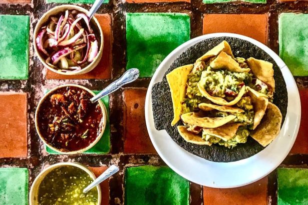 Guacamole servi dans un molcajete de pierre avec des chips de tortilla, trois salsas et des oignons marinés, sur un carrelage mexicain vert et terracotta